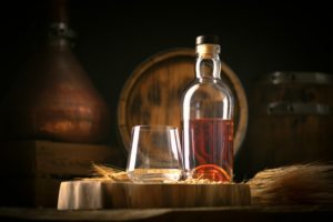 A clear bottle filled halfway with whisky, sitting on a barrel next to a rocks glass in front of a stack of other brown barrels