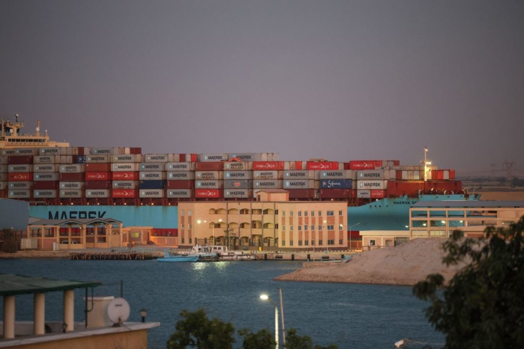 A HUGE CONTAINER SHIP PUSHES THROUGH A NARROW WATERWAY, BUILDINGS VISIBLE IN THE FOREGROUND