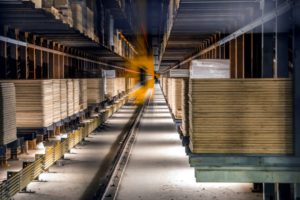 MANY SHEETS OF CONSTRUCTION MATERIAL ARE STACKED IN A WAREHOUSE, A BLURRED YELLOW ROBOT MOVING DOWN THE AISLE BETWEEN THEM